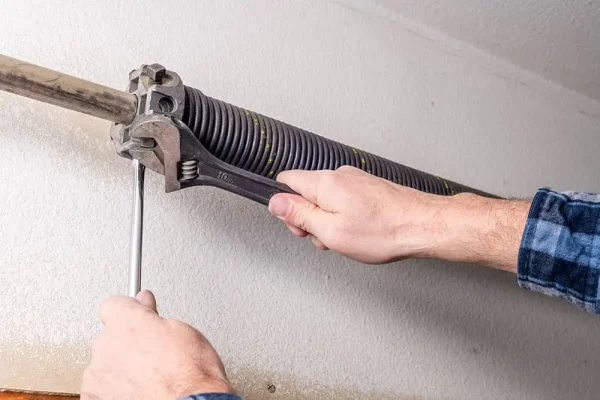 A close-up of a person using a wrench and hex key to adjust or repair a torsion spring on a garage door.